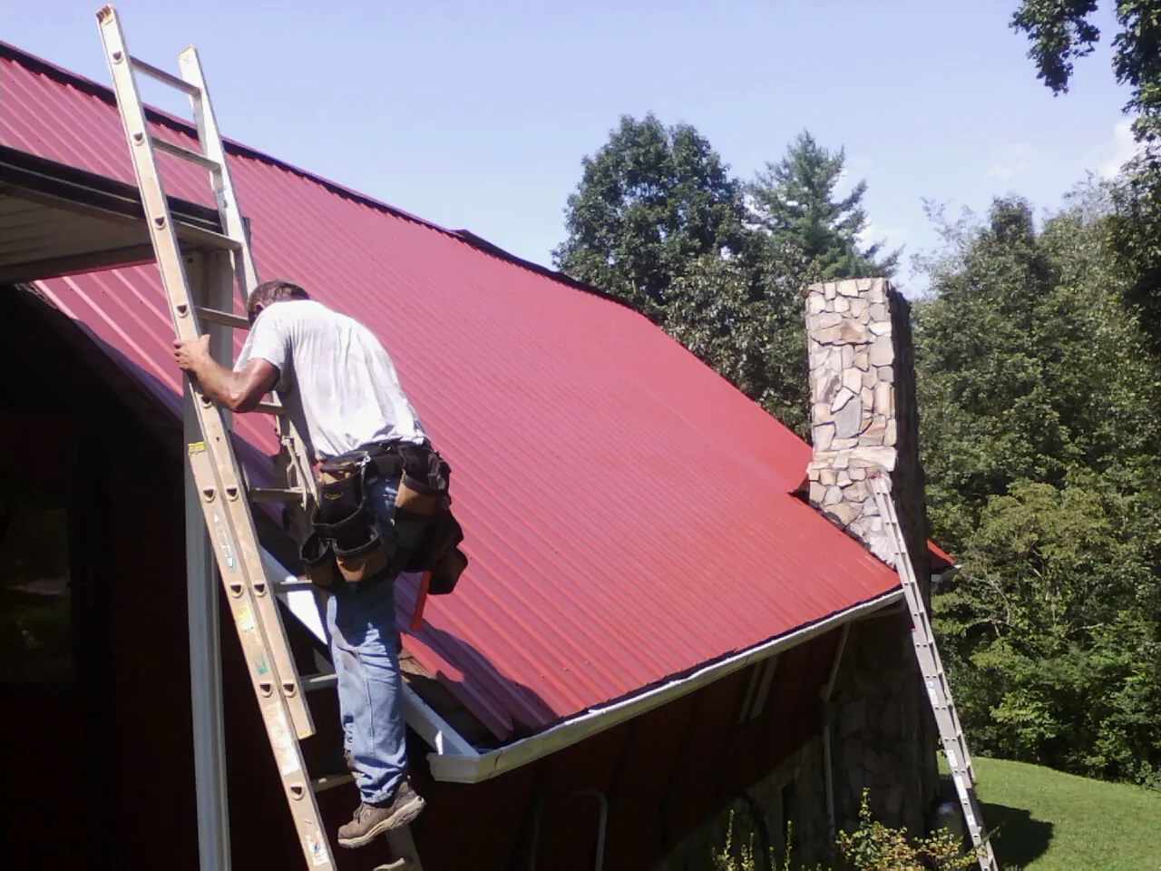 Roofer on a ladder beside a red metal roof and stone chimney — Soffit & Fascia Repair by Roofing Brothers in Piketon